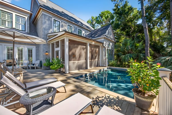 Modern coastal home with pool, lounge chairs, screened porch, and mature trees surrounding the backyard patio area.